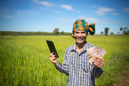 Happy Asian man farmer with smiling face hand holding smart phone standing in rice farm, cash subsidy conceptの写真素材