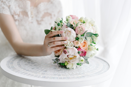 Bride touching wedding bouquet lying on white table with one hand with wedding ring on her fingerの写真素材