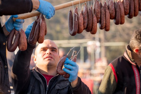 PERNIK, BULGARIA - JANUARY 26, 2018: Bald man with blue latex gloves arranges several home-made pork sausages to dry and to be shown at the annual International Festival of Masquerade Games Survaのeditorial素材