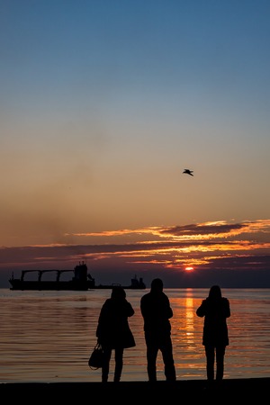 The persons silhouettes standing by the sea and enjoying the scenery view at sunset with water reflections and ship, golden hour calmnessの写真素材