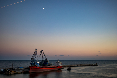BALCHIK, BULGARIA - SEPTEMBER 17, 2018: Middle sized red transport ship in calm sea waters gets loaded at Black Sea port by two cranes at summer evening in blue hour, colorful waterscapeのeditorial素材