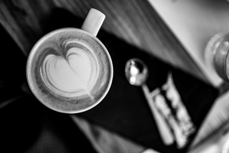 Selective shallow focus, view directly from above on the foam in a shape of heart in a cup of cappuccino. Spoon on black napkin and pack of sugar on a table out of focus. Black and white impressionの写真素材