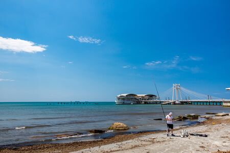 DURRES, ALBANIA - MAY 17, 2017: Young fisherman fixes his fishing equipment on the city beach in sunny blue sky spring day. The modern architecture Ventus Harbour is in the backgroundのeditorial素材