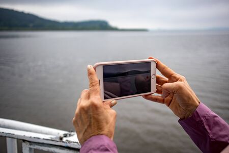 Woman hands with pink sleeves holding smart phone shooting video of lake Pamvotida near the Greek town of Ioannina, early spring morning with fog, selective focusの写真素材