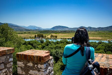 High angle sunny spring day view of salt lagoon lake, valley and mountain in the national park of Butrint in Albania, Europe. Blurred figure of female photographer and fort walls in the foreground.の写真素材