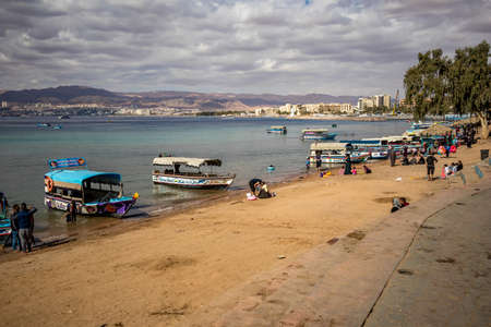 AQABA, JORDAN - JANUARY 31, 2020: Local people and tourists enjoy holiday Friday at sunny city beach with glass floor boats. Winter puffy clouds morning sky. Red Sea gulf, Hashemite Kingdom of Jordanのeditorial素材