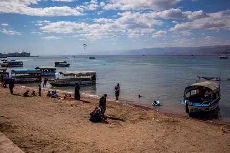 AQABA, JORDAN - JANUARY 31, 2020: Local people and tourists enjoy holiday Friday at sunny city beach with glass floor boats. Winter puffy clouds morning sky. Red Sea gulf, Hashemite Kingdom of Jordanのeditorial素材