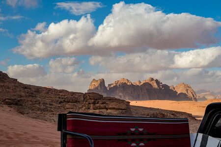 Jordan, Wadi Rum desert, winter day landscape with white puffy clouds. Beautiful desert could be explored on safari. Blurred bench in foreground on pickup SUV truck covered with rug for tourists.の写真素材