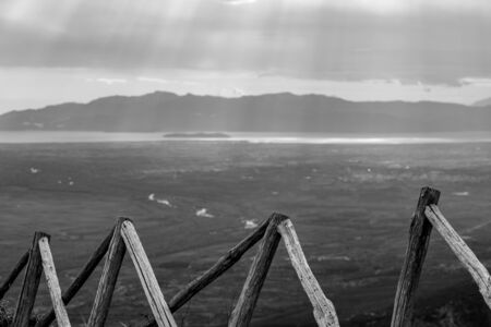 Wooden fence high in mountains with blurred sun rays through heavy clouds over sea in Northern Greece. High angle view from the mountains at the Greek region of Xanthi. Black and white travel photoの写真素材