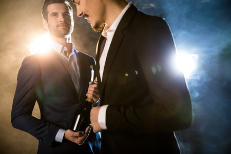 Two stylish men in suits with champagne bottle smiling under spotlightsの写真素材