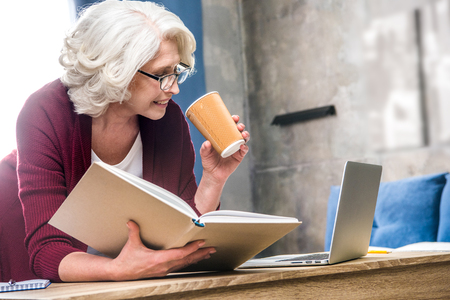 Attractive senior woman holding paper cup and reading bookの写真素材