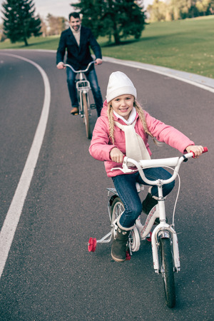 Cute smiling little girl cycling with father on road in autumn parkの写真素材