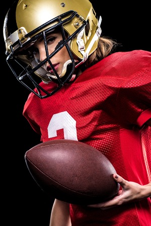 Beautiful female american football player in sportswear and helmet holding ball on blackの写真素材