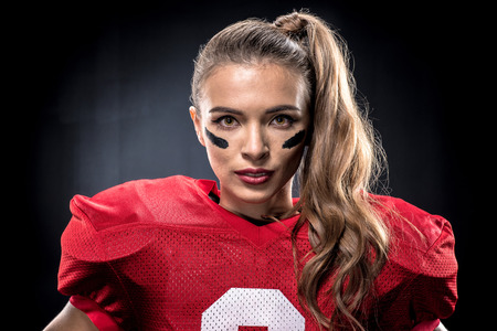 Close-up portrait of beautiful female american football player in uniform posing and looking at camera on blackの写真素材