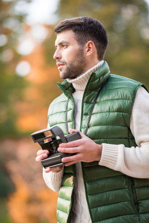 Handsome young man holding instant camera and looking away outdoors
