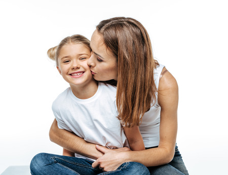 Close-up portrait of mother hugging and kissing cute smiling daughter isolated on whiteの写真素材
