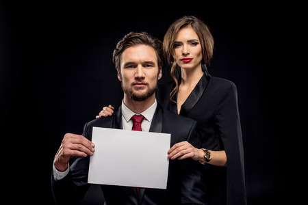 Beautiful couple in formal wear holding blank white card and looking at camera on blackの写真素材