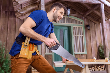 smiling bearded carpenter in tool belt sawing wooden plank on porchの写真素材