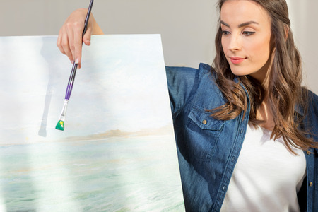 young woman artist with paintbrush leaning at easel with pictureの写真素材