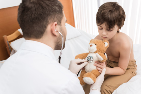 doctor with stethoscope examining little boy holding teddy bearの写真素材