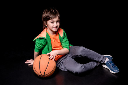 smiling boy sitting on floor with basketball ball and looking awayの写真素材