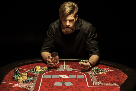 man with glass of alcohol drink and poker chip in hands sitting at poker tableの写真素材