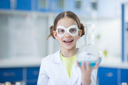 girl scientist in protective glasses holding flask and smiling at cameraの写真素材
