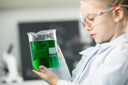 girl wearing lab coat and protective glasses holding flask with green reagent