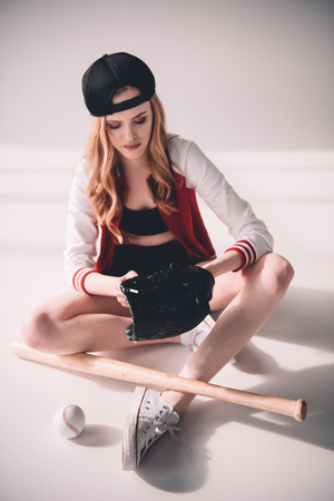 young sporty woman in cap sitting with baseball equipmentの写真素材