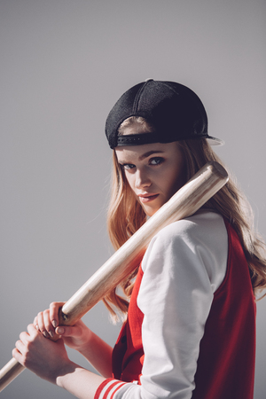 young woman in cap holding baseball bat and looking at cameraの写真素材