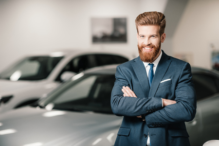 salesman in suit with crossed arms posing and looking at camera in dealership salonの写真素材
