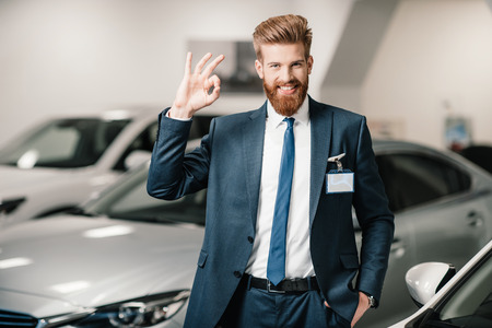 salesman in suit showing ok sign and looking at camera in dealership salonの写真素材