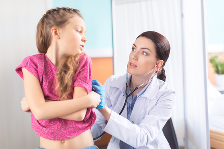 woman doctor with stethoscope listening to heartbeat and lungs of little girl patientの写真素材