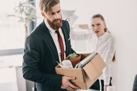 upset fired businessman with cardboard box and angry businesswoman in officeの写真素材