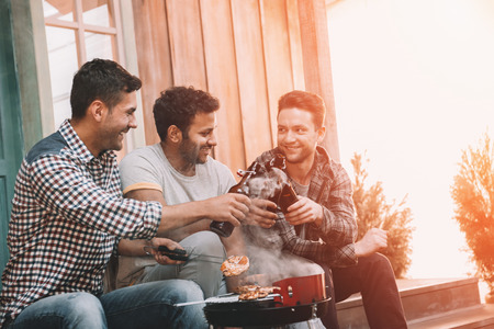 young men clinking beer bottles and grilling meat on porchの写真素材