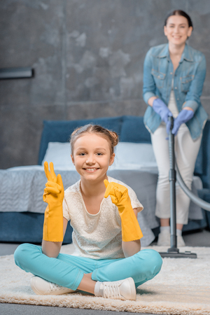Mother with vacuum cleaner looking at happy daughter in rubber gloves gesturingの写真素材