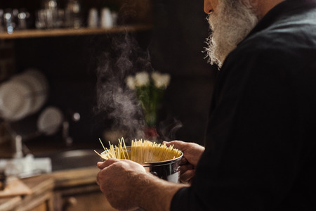 Partial view of bearded senior man holding pan with spaghetti in kitchenの写真素材