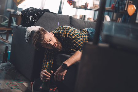 Drunk bearded young man holding beer bottle lying on couch in messy roomの写真素材