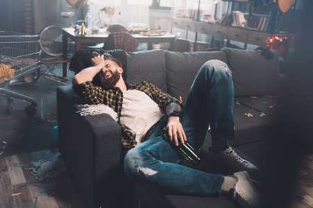 bearded young man with bottle lying on sofa after partyの写真素材