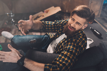 bearded young man with hangover gesturing in messy room after partyの写真素材