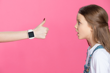 shocked little girl looking at woman with smartwatch showing thumb upの写真素材