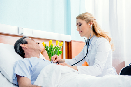 middle aged patient lying on bed and doctor examining him with stethoscopeの写真素材