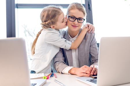mother and adorable daughter hugging and kissing in office with laptopsの写真素材
