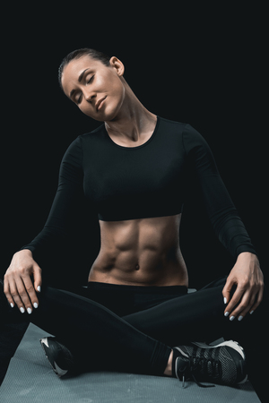 fitness woman stretching neck while sitting on mat isolated on black.の写真素材