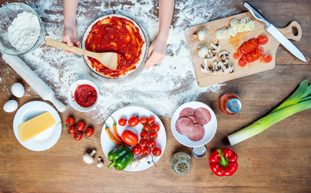 child making pizza with pizza ingredients, tomatoes, salami and mushrooms on wooden tabletopの写真素材