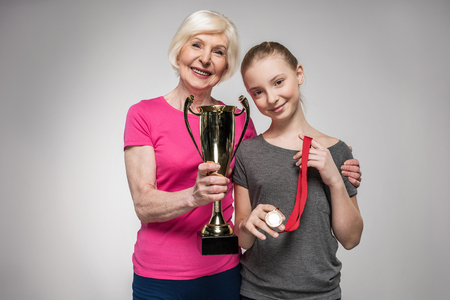 senior sportswoman and girl holding trophy and medal isolated on greyの写真素材