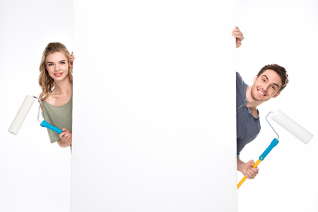 Smiling young couple with paint rollers and blank card looking at cameraの写真素材