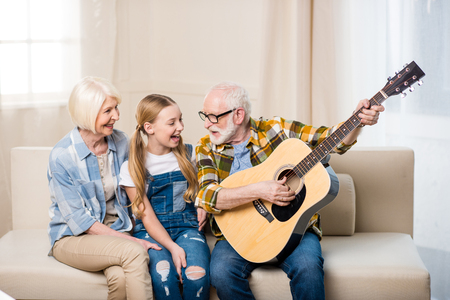 happy girl with grandparents sitting together on sofa and playing acoustic guitarの写真素材
