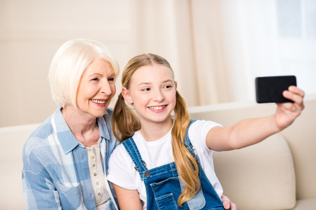 grandmother and granddaughter sitting together on sofa and taking selfie with smartphoneの写真素材