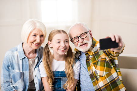 girl with grandmother and grandfather sitting on sofa and taking selfieの写真素材